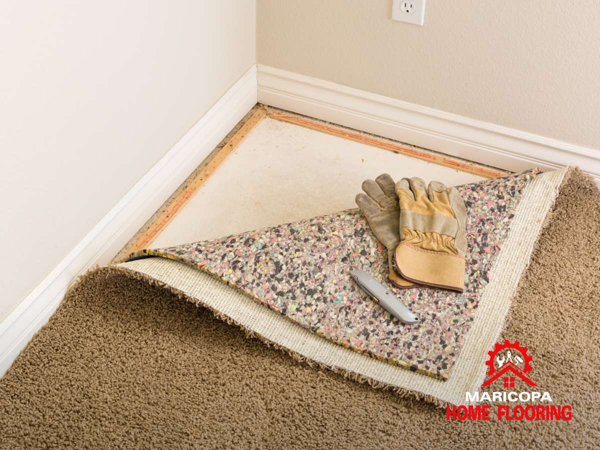 Carpet removal in a room corner with padding, gloves, and utility knife on subfloor.